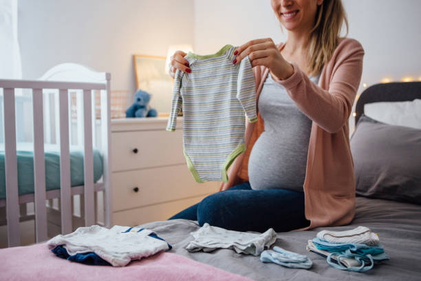 Pregnant young woman holding baby clothing, sitting on bed next to the crib and smiling